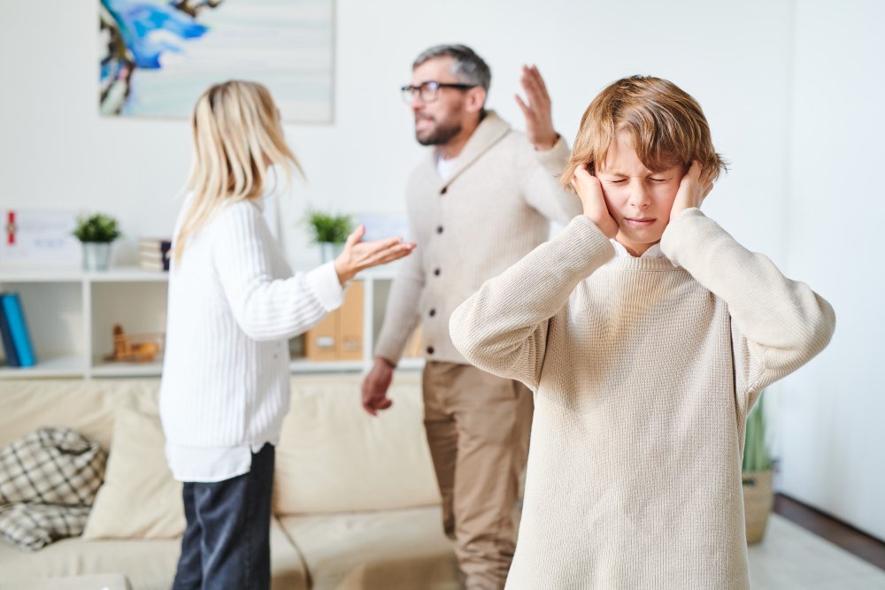 Child covering ears in distress while parents argue in the background, a common cause of childhood anxiety disorder